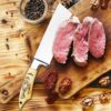 Handmade chef cleaver slicing cooked beef on wooden cutting board.