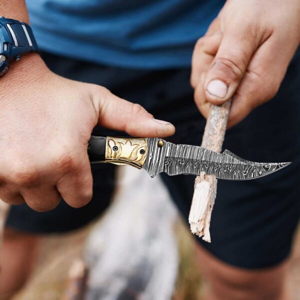 Handmade Damascus folding knife carving wood during outdoor camping activity.